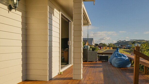A wooden deck with a bench and a blue bean bag chair, a white building with a sliding door, and a clear sky. - Home Base - Cooks Beach Holiday Home (Cooks Beach)