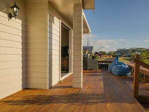 Una terraza de madera con un banco y un sillón informal de cuentas azul, un edificio blanco con puerta corredera, y un cielo despejado.