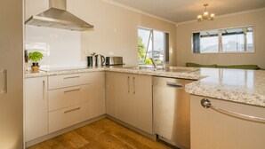 A modern kitchen with white cabinets, a stainless steel range hood, a dishwasher, and a granite countertop. - Home Base - Cooks Beach Holiday Home (Cooks Beach)