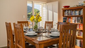 A dining area with a wooden table set for four, surrounded by chairs. A bookshelf filled with books is to the right. - Home Base - Cooks Beach Holiday Home (Cooks Beach)