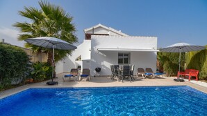 A swimming pool with lounge chairs and umbrellas, surrounded by greenery and a white building.