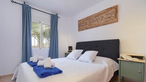 A neatly made bed with white and blue linens, a bedside table with a phone and a book, and a window with blue curtains.