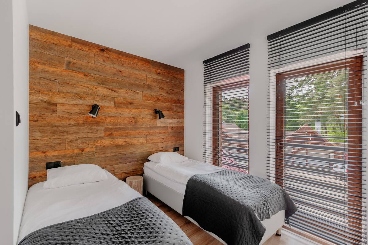 A bedroom with two beds, wooden wall paneling, and large windows with blinds.