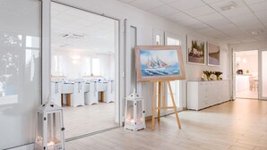 Een ruime kamer met een groot schilderij op ezel, witte meubels en uitzicht op een andere kamer met daarop opgestelde tafels.