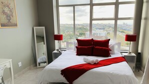 Ein Hotelzimmer mit einem großen Bett, roten Kissen, weißer Bettwäsche und Blick auf die Stadt durch große Fenster.
