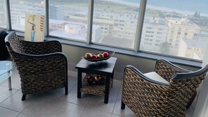 Ein Zimmer mit Korbstühlen, einem kleinen Tisch mit Dekoration und Blick auf das Meer sowie Gebäude durch große Fenster.