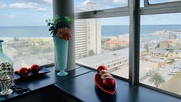 Blick aus einem Fenster auf eine Skyline mit Strand, einem hohen Gebäude und einem Blumentopf auf der Fensterbank.