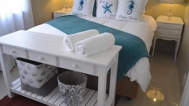 Quarto de hotel bem organizado, com cama, mesas de cabeceira, uma pequena mesa com toalhas, um balde estampado e uma caneca decorativa.