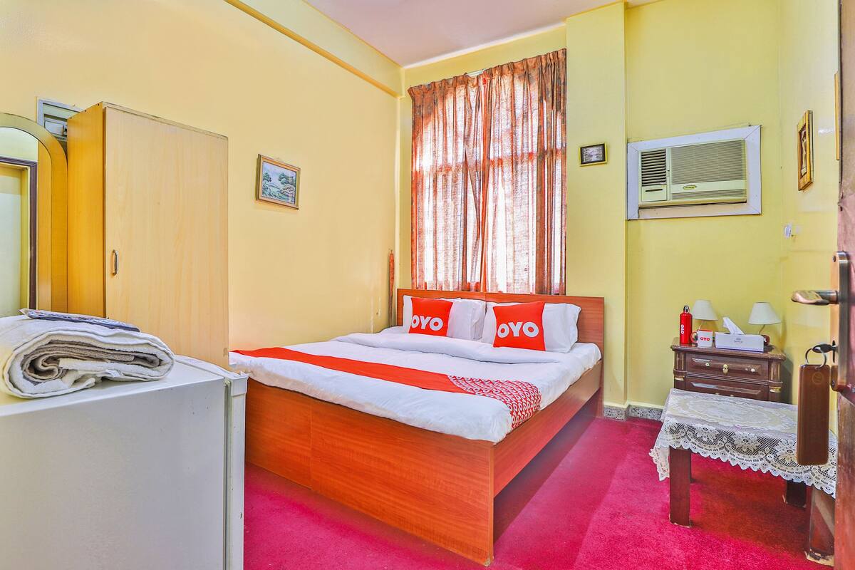 A bedroom with a wooden bed, a small bedside table, a nightstand, a wardrobe, a window with curtains, and a wall-mounted air conditioner.