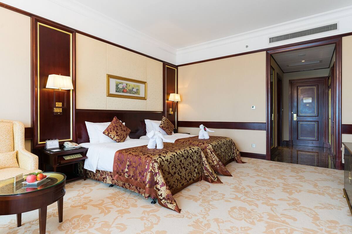A hotel room with two beds, a sitting area with a chair, a small table with a fruit bowl, and a painting on the wall.