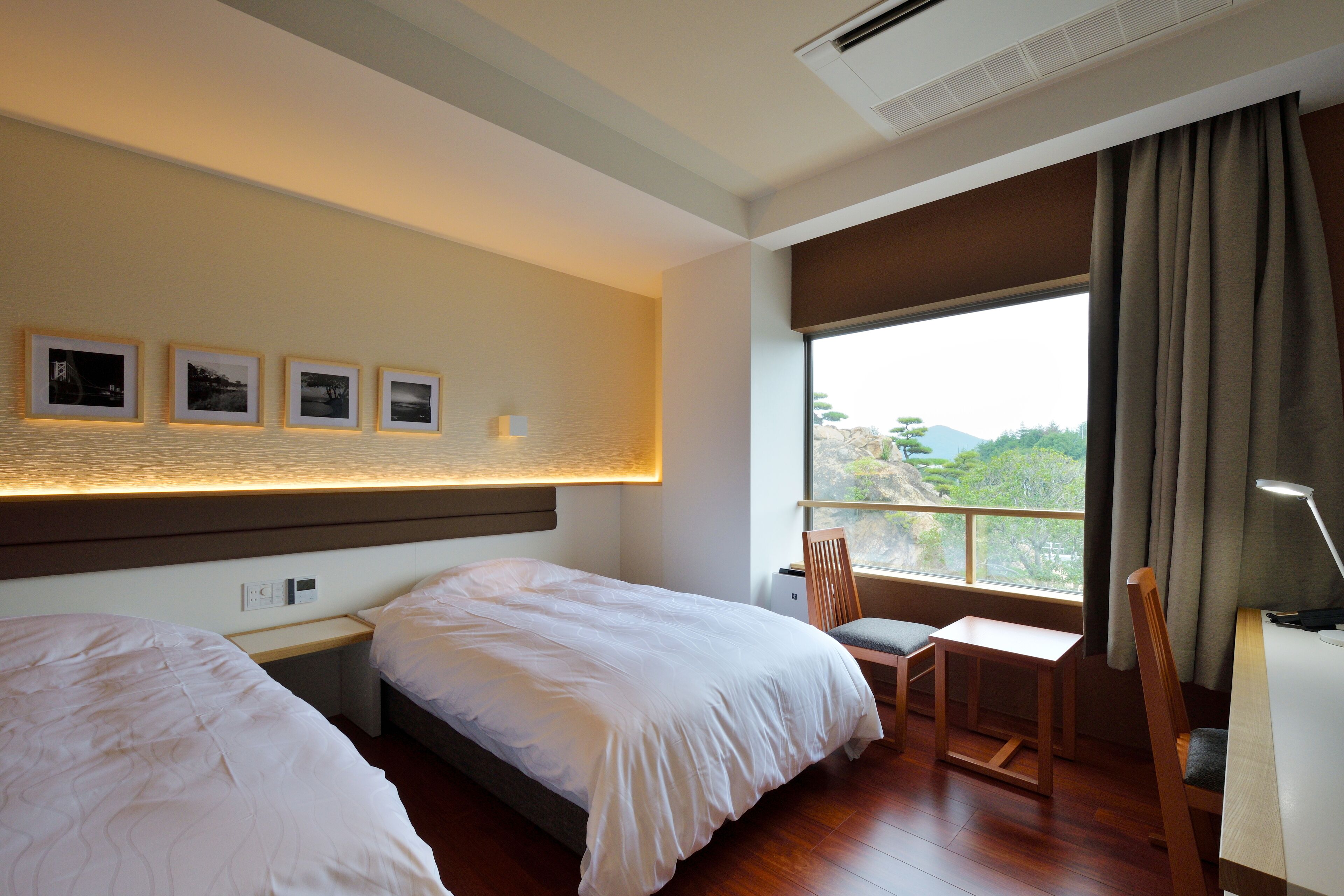 Habitación de hotel con dos camas, un escritorio, una silla y un amplio ventanal con cortinas.