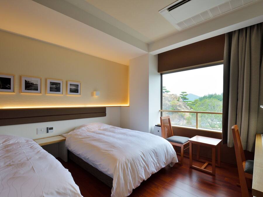 Habitación de hotel con dos camas, un escritorio, una silla y un amplio ventanal con cortinas.