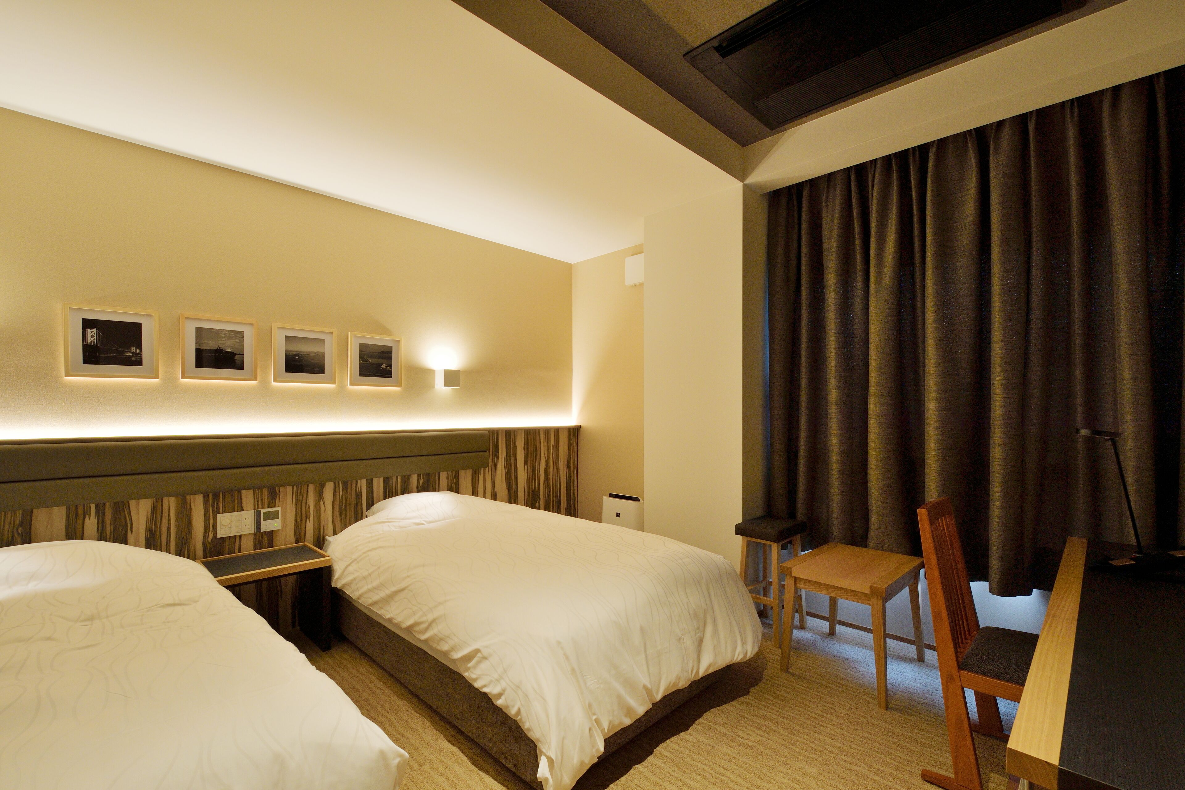 A hotel room with two beds, a desk, a chair, and a wall-mounted light fixture.
