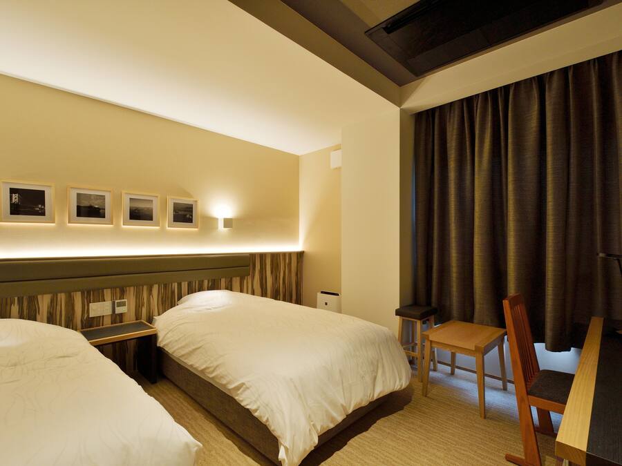 A hotel room with two beds, a desk, a chair, and a wall-mounted light fixture.