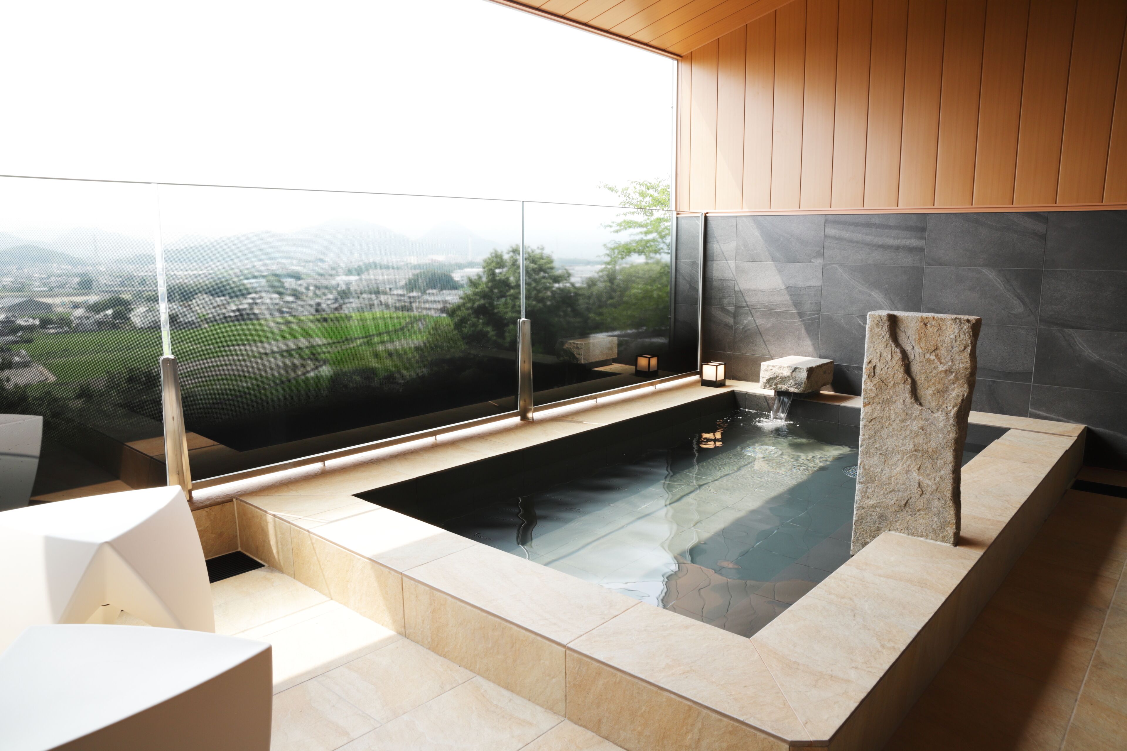 A modern spa with a glass-enclosed hot tub, a stone pillar, and a view of a green landscape.