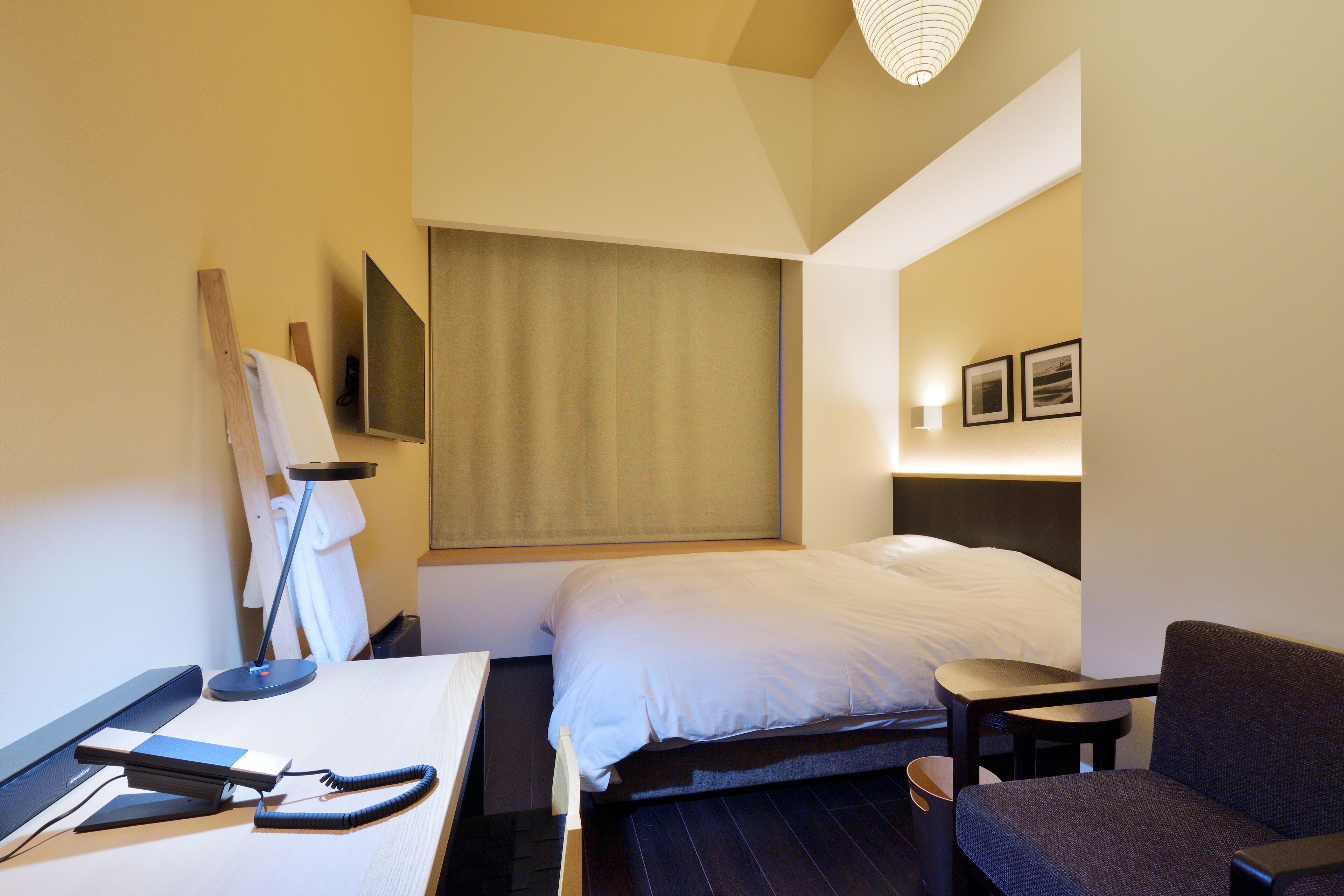 A hotel room with a bed, a desk with a computer, a chair, and a bedside table.