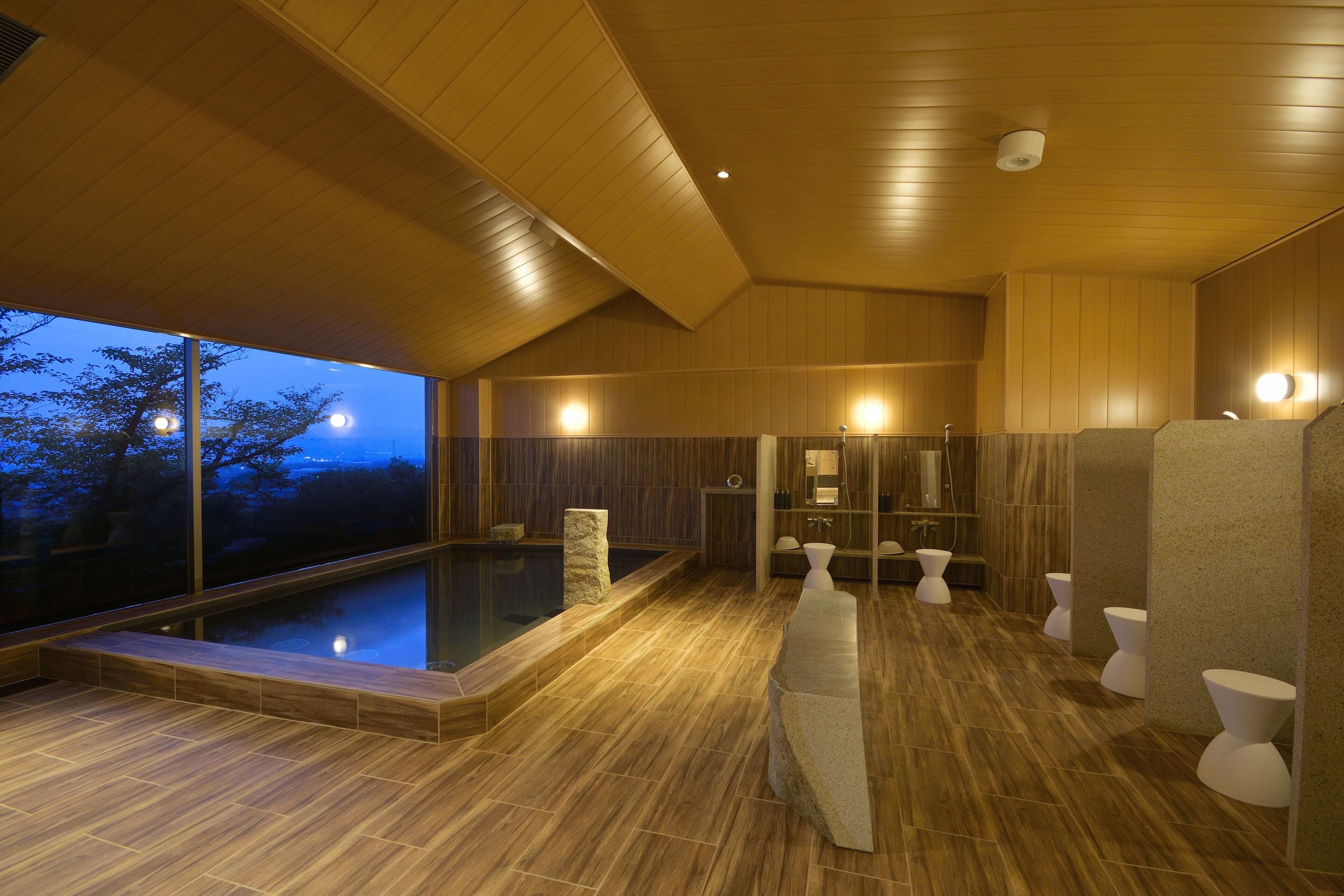 A modern indoor pool area with a wooden ceiling and floor, stone benches, and a view of the outdoors at dusk.