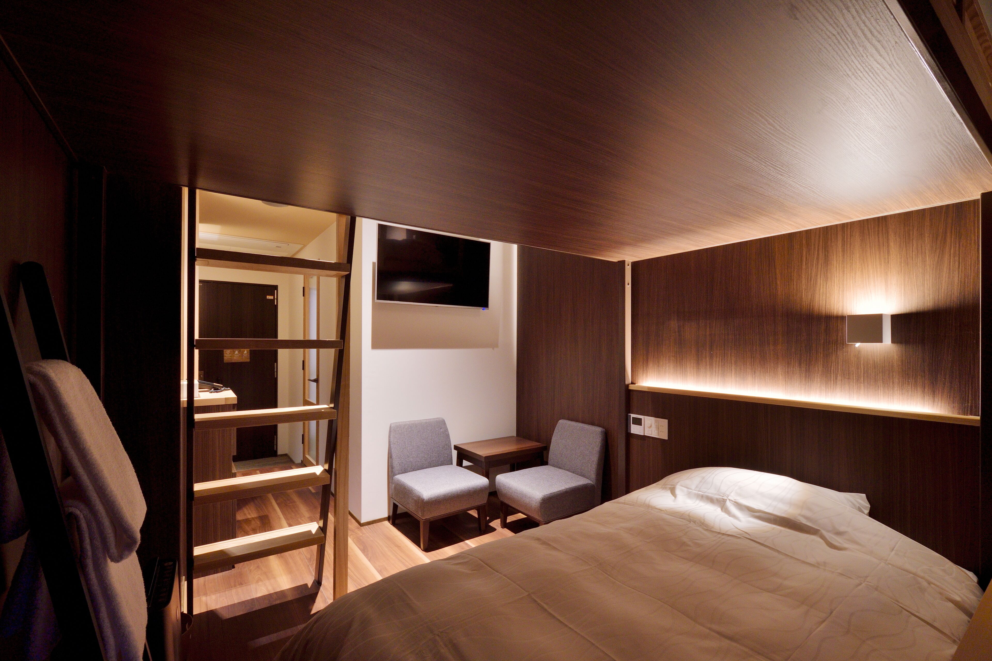 A modern hotel room with a bed, two chairs, a small table, a TV, and a built-in shelf.