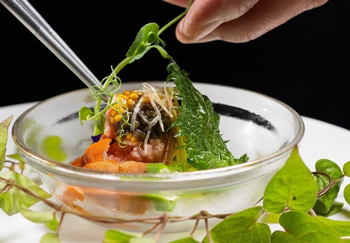 Un tazón de vidrio transparente con diversos alimentos, incluyendo aparentemente maíz, vegetales de hoja verde y posiblemente carne, decorado con una hoja.