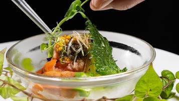 Un tazón de vidrio transparente con diversos alimentos, incluyendo aparentemente maíz, vegetales de hoja verde y posiblemente carne, decorado con una hoja.