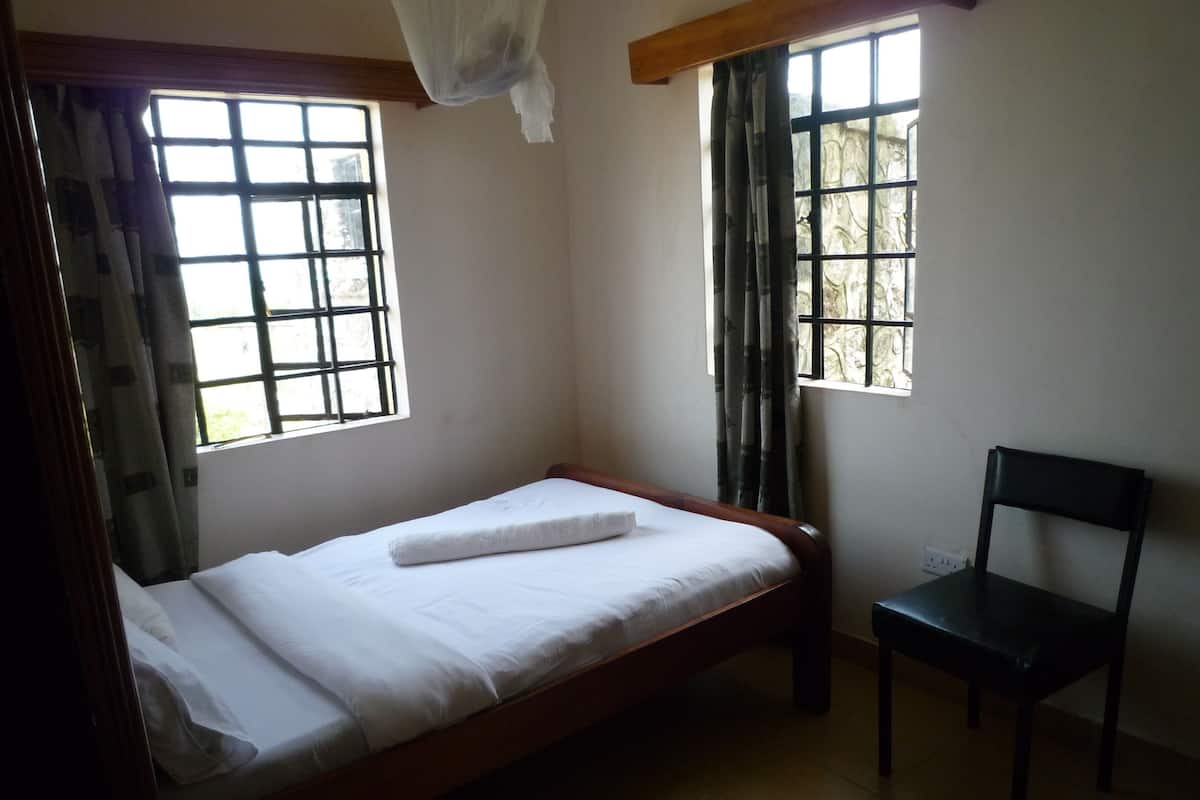 A single bed with a white mattress and pillows, a wooden headboard, a chair, and two windows with curtains.