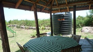 A covered outdoor dining area with a table set for four, surrounded by wooden chairs. A large black water tank labeled "POA TANK" is visible in the background. - Cawaal Hotels (Kisumu)
