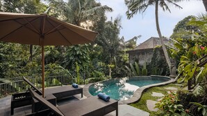 A poolside area with lounge chairs and an umbrella, surrounded by lush greenery and a stone building. - Andara Ubud Villa (Ubud)