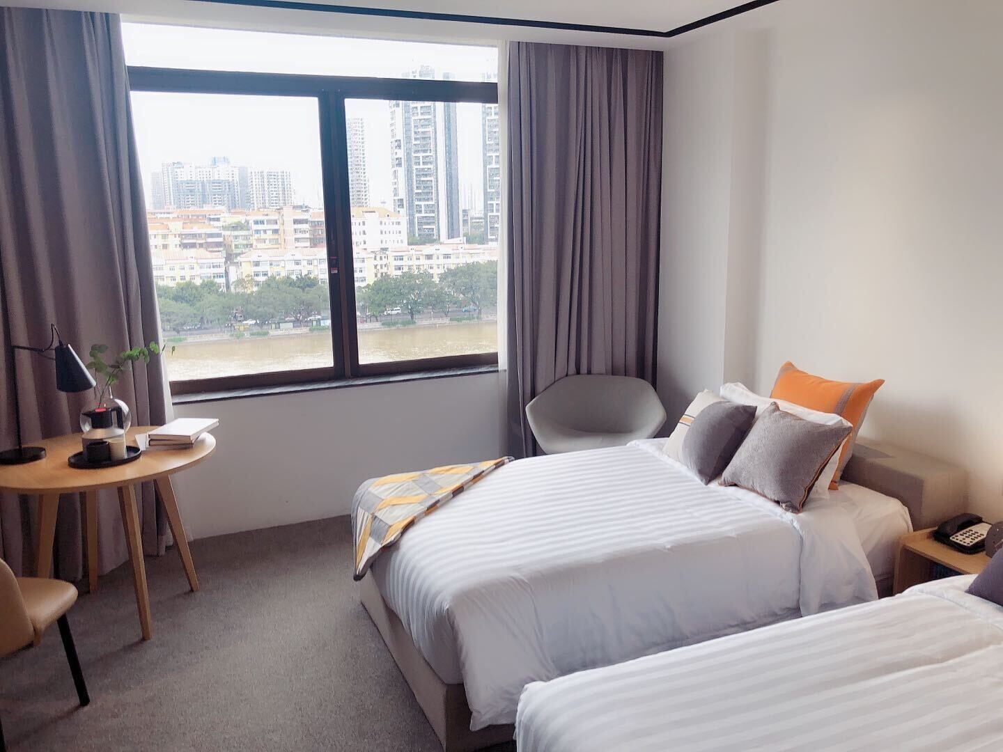 A hotel room with a large window, a desk with a lamp, two beds, and a chair.