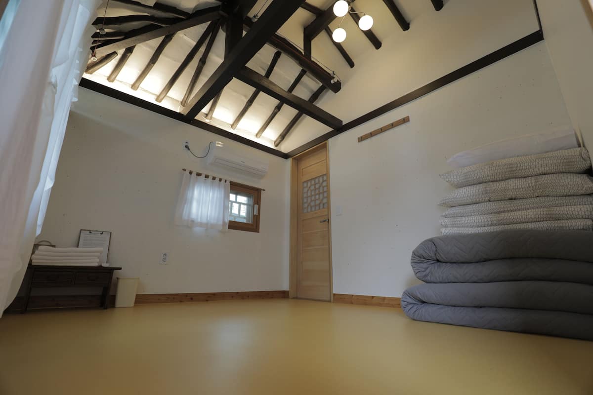 Une pièce avec un plafond en pente, un banc en bois avec des serviettes, une fenêtre avec des rideaux et une pile d’oreillers.