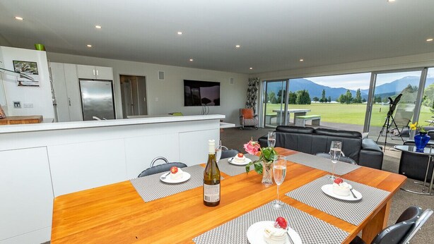 Una cocina moderna con gabinetes blancos y electrodomésticos de acero inoxidable, un comedor con una mesa de madera dispuesta para cuatro personas, y un amplio ventanal con vistas al exterior.