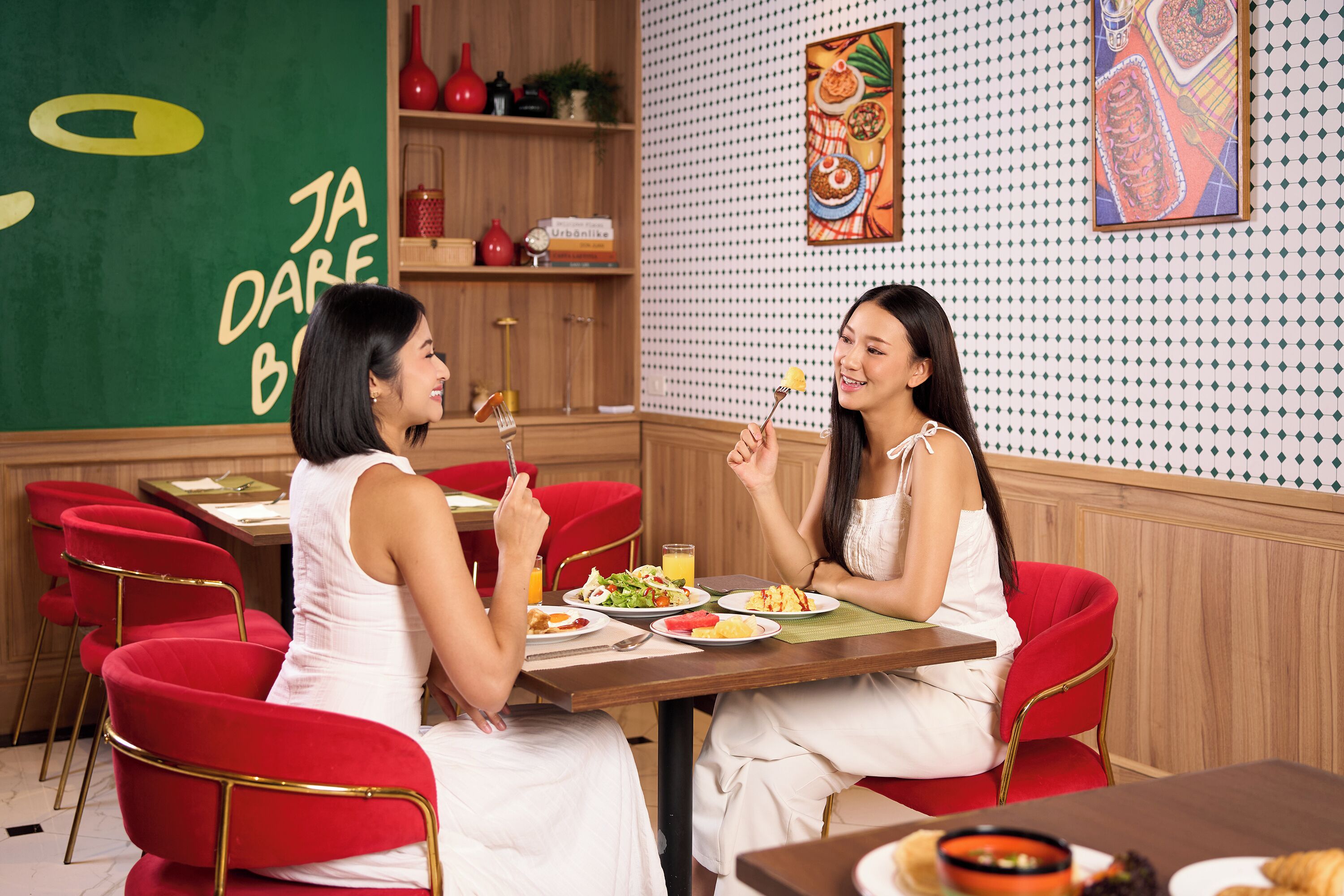 Two individuals seated at a table with various dishes, in a room with a green wall and a patterned wallpaper.