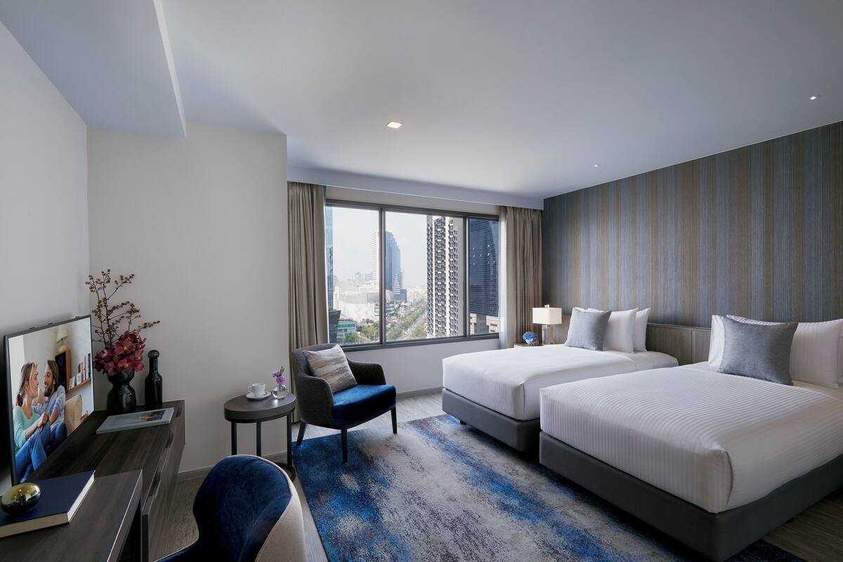 Une chambre d’hôtel avec deux lits, un bureau avec un ordinateur, une chaise, une petite table avec une lampe et une grande fenêtre donnant sur la ville.