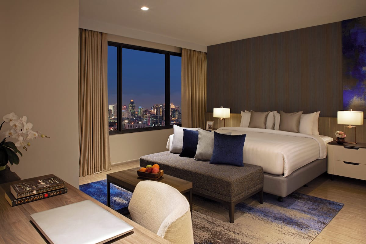 Une chambre d’hôtel moderne avec un grand lit, un bureau avec un ordinateur portable, une table basse et une vue sur le paysage urbain au crépuscule.