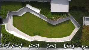 An aerial view of a landscaped area with a central grassy lawn, surrounded by concrete borders and bordered by dense greenery.