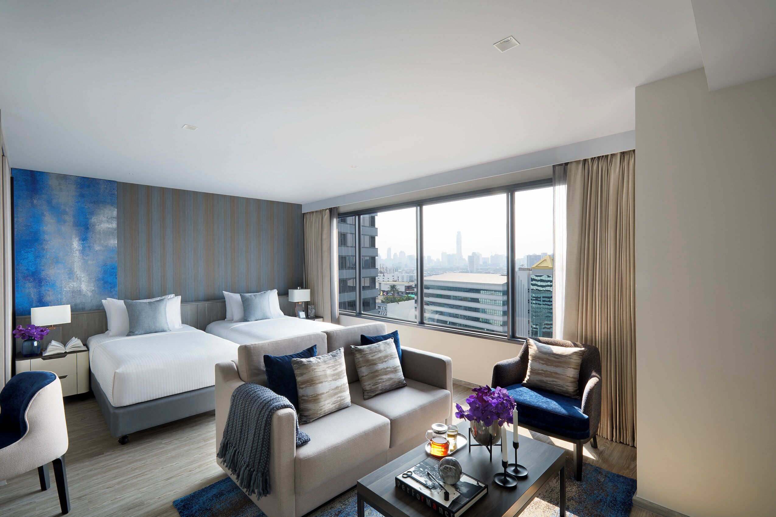 Quarto de hotel moderno com duas camas, um sofá, uma mesa de centro e vista para o horizonte da cidade.
