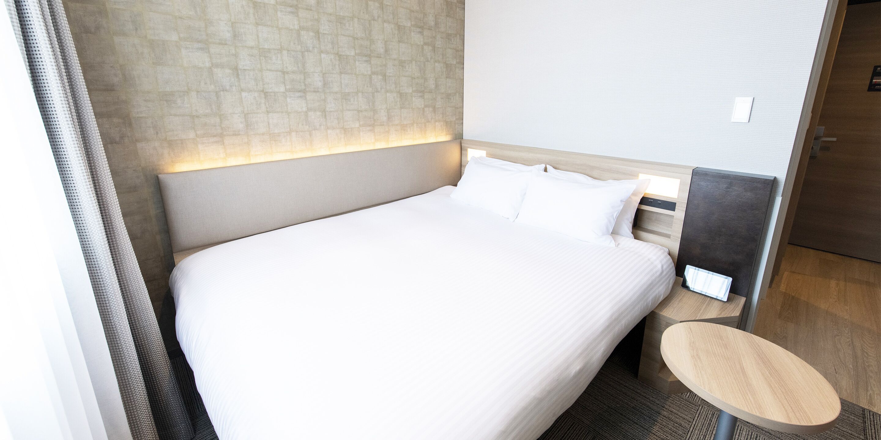 A hotel room with a large bed, a bedside table, and a headboard with a light panel.