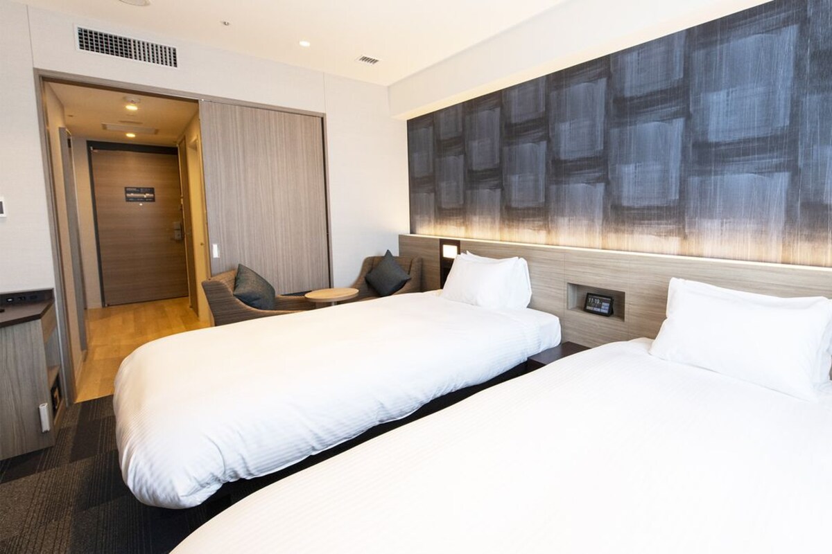 Ein Hotelzimmer mit zwei Betten, einem großen, abstrakten Gemälde an der Wand, einer Holztür und einem kleinen Tisch mit einer Lampe.