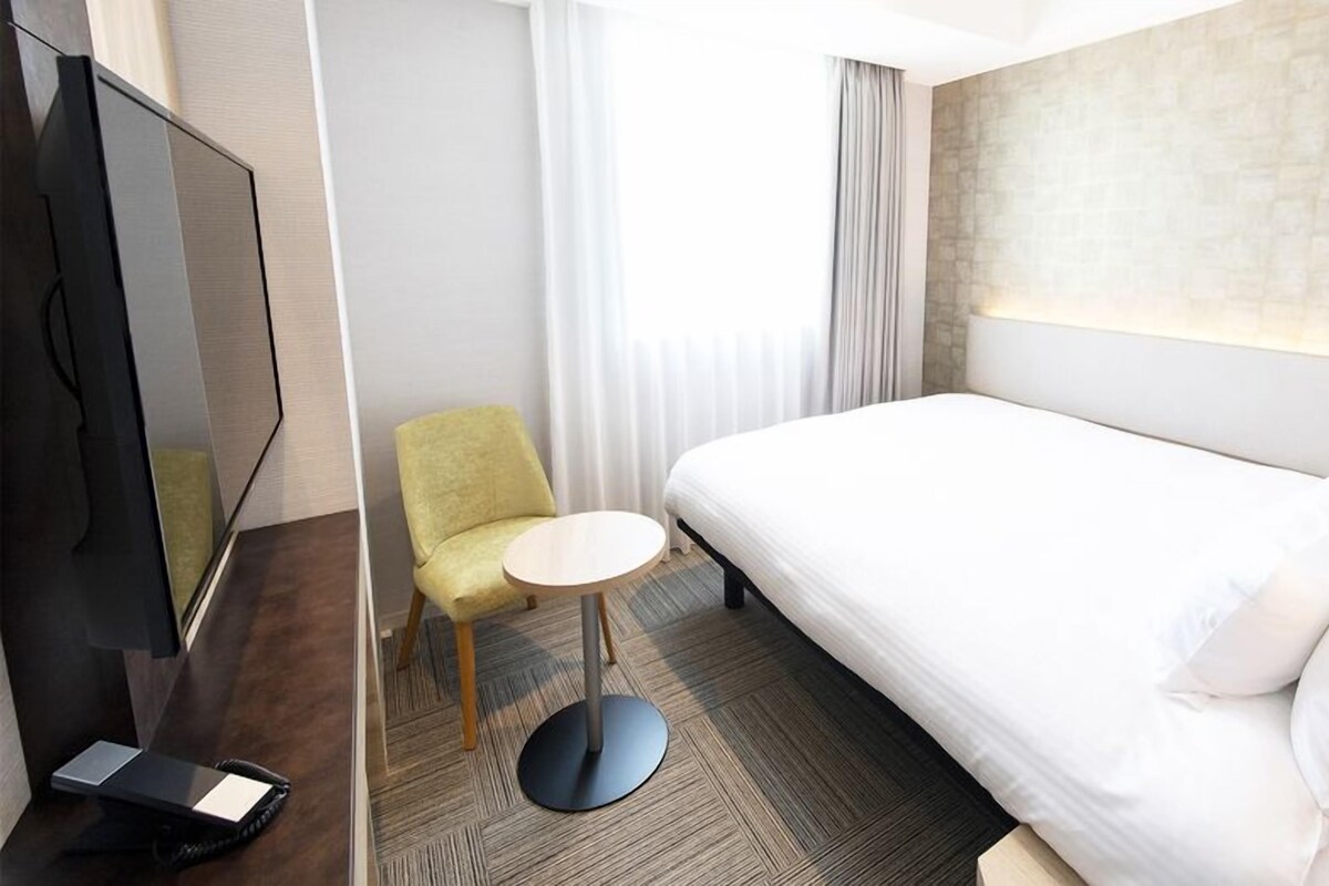 Ein Hotelzimmer mit Bett, kleinem runden Tisch, Sessel, Fernseher und Fenster mit Vorhängen.