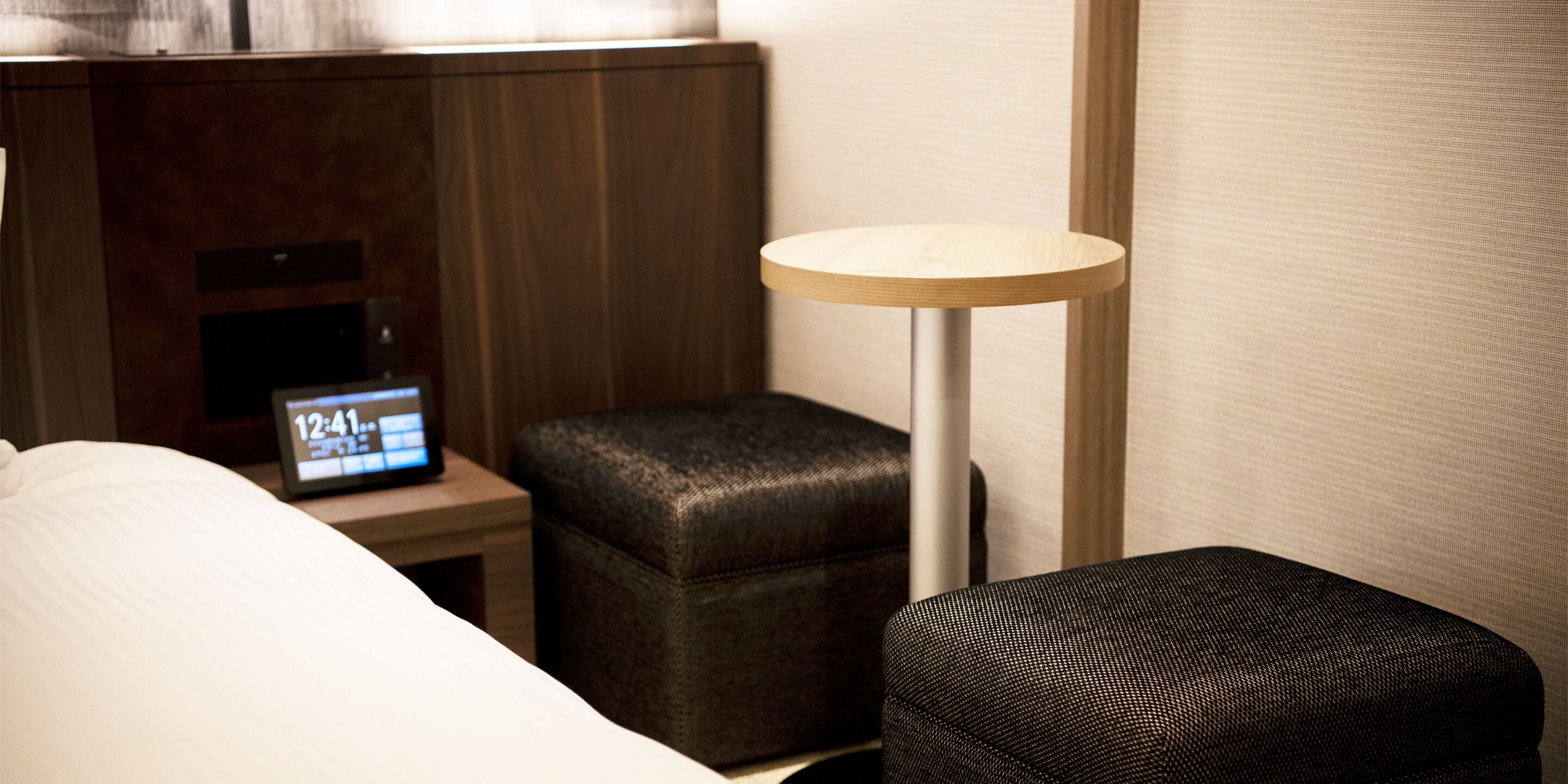 A hotel room with a bed, a bedside table with a clock, a small ottoman, and a floor lamp.