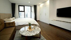 Une chambre d’hôtel moderne avec un lit, une table basse ronde sur laquelle se trouvent un ananas et une tasse, un canapé marron, un téléviseur fixé au mur et une vue sur la ville par la fenêtre.