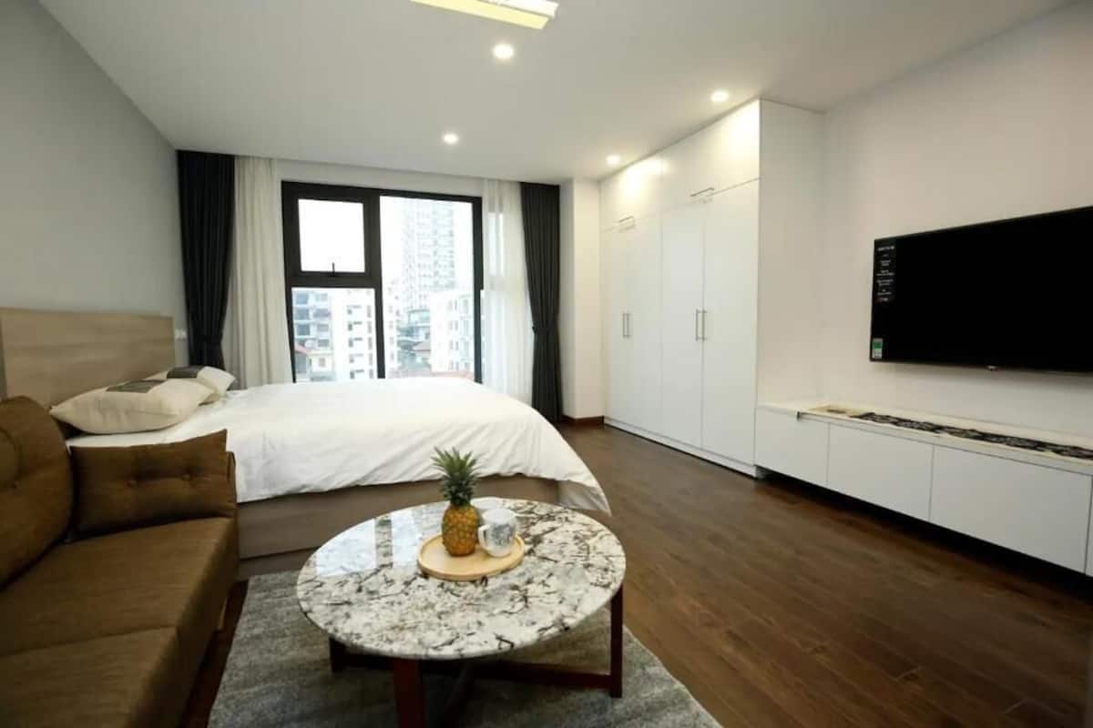 Ein modernes Hotelzimmer mit einem Bett, einem runden Couchtisch mit Ananas und Tasse, einer braunen Couch, einem Fernseher an der Wand und Blick auf die Stadt durch das Fenster.