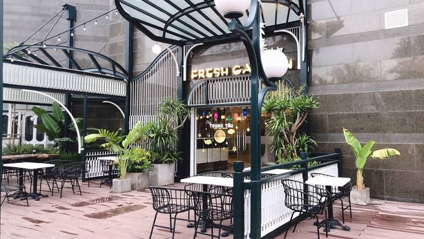 Un café en plein air avec des tables et des chaises, une enseigne indiquant « CAFÉ FRAIS » et des plantes en pot.