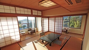 A tatami mat room with a low table and chairs, large windows with a view of a mountain and buildings, and traditional sliding doors. - Kashikojima Hotel Bay Garden (Shima)