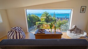A room with a large window offering a view of the sea and mountains, a wooden table with a bottle and glasses, and a bean bag chair.