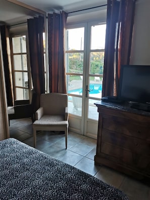 Une chambre comprenant un lit, une chaise, un téléviseur, une armoire en bois et une porte-fenêtre coulissante donnant sur une zone avec piscine.