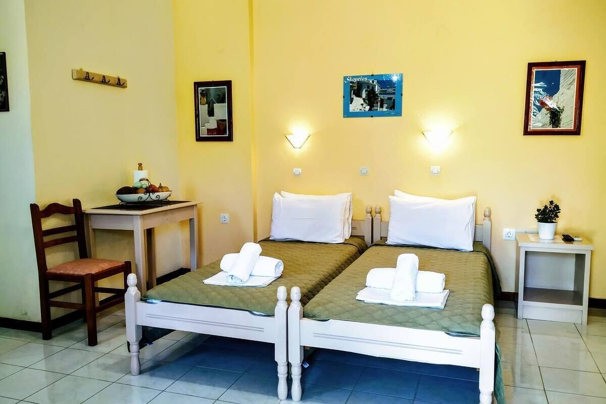 A double bed with white bedding and towels, a small table with a bowl of fruit, a chair, and a bedside table with a plant.