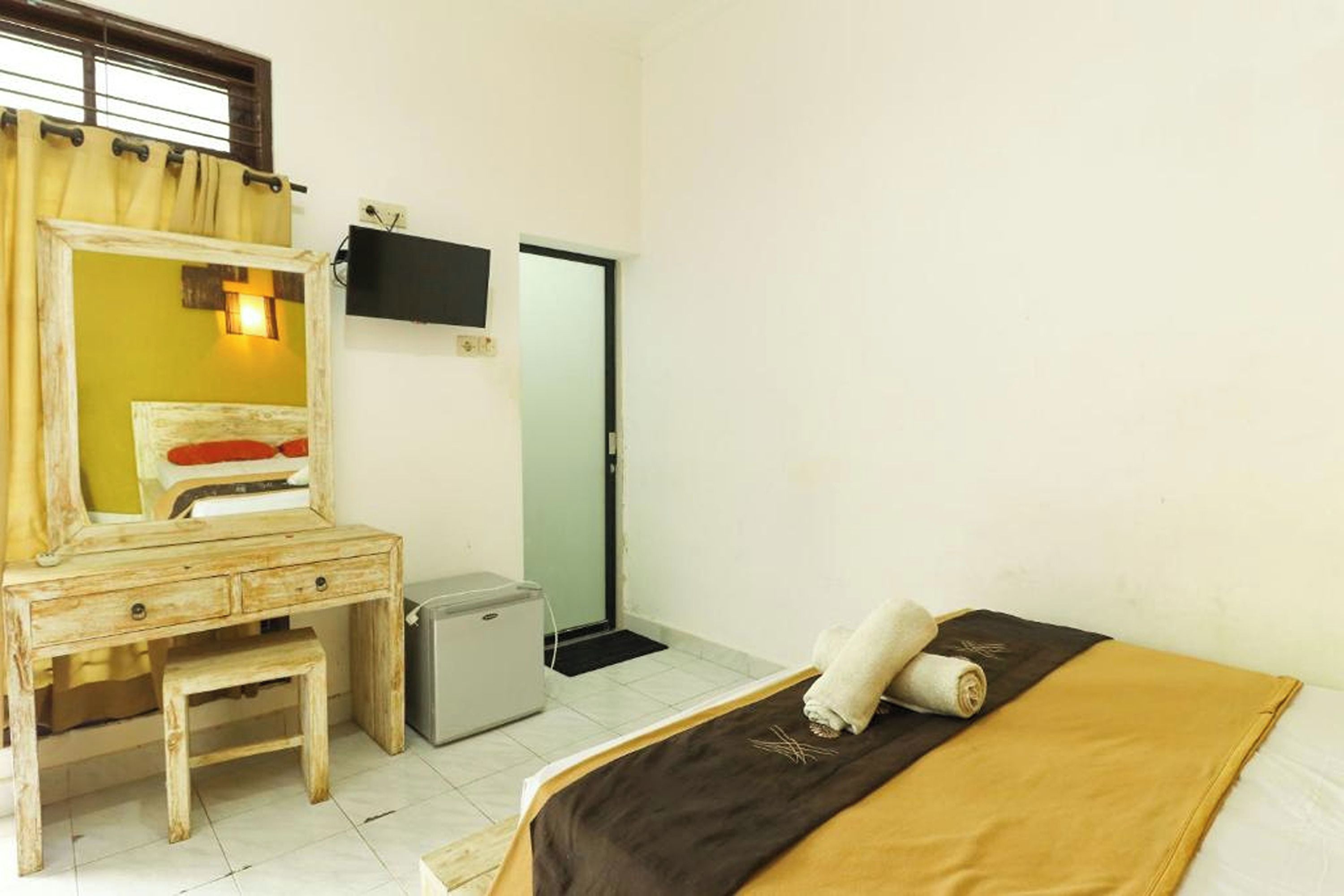 A room with a bed, a small desk with a mirror, a television mounted on the wall, and a door.