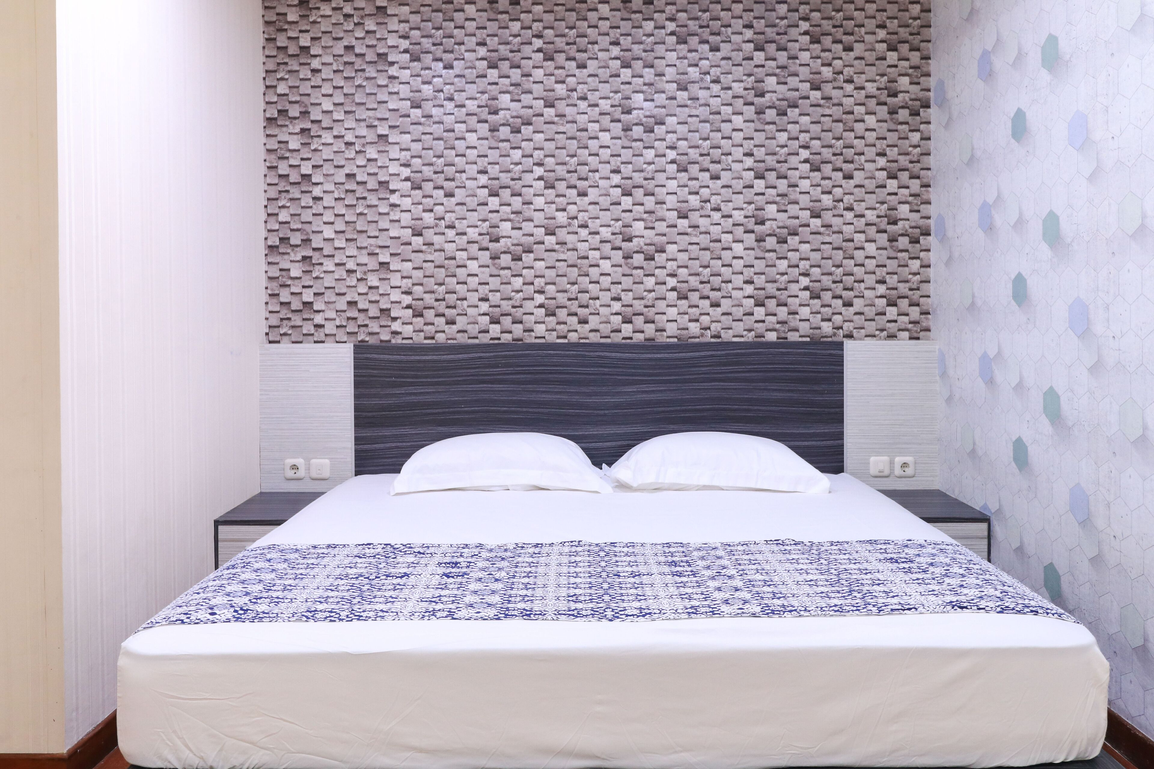 A neatly made bed with a patterned bedspread, a headboard with a textured wall behind it, and a wall with a geometric pattern.