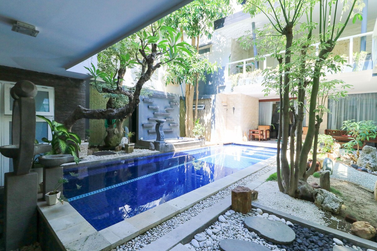 Una piscina con borde de piedra y guijarros, rodeada de árboles y plantas.