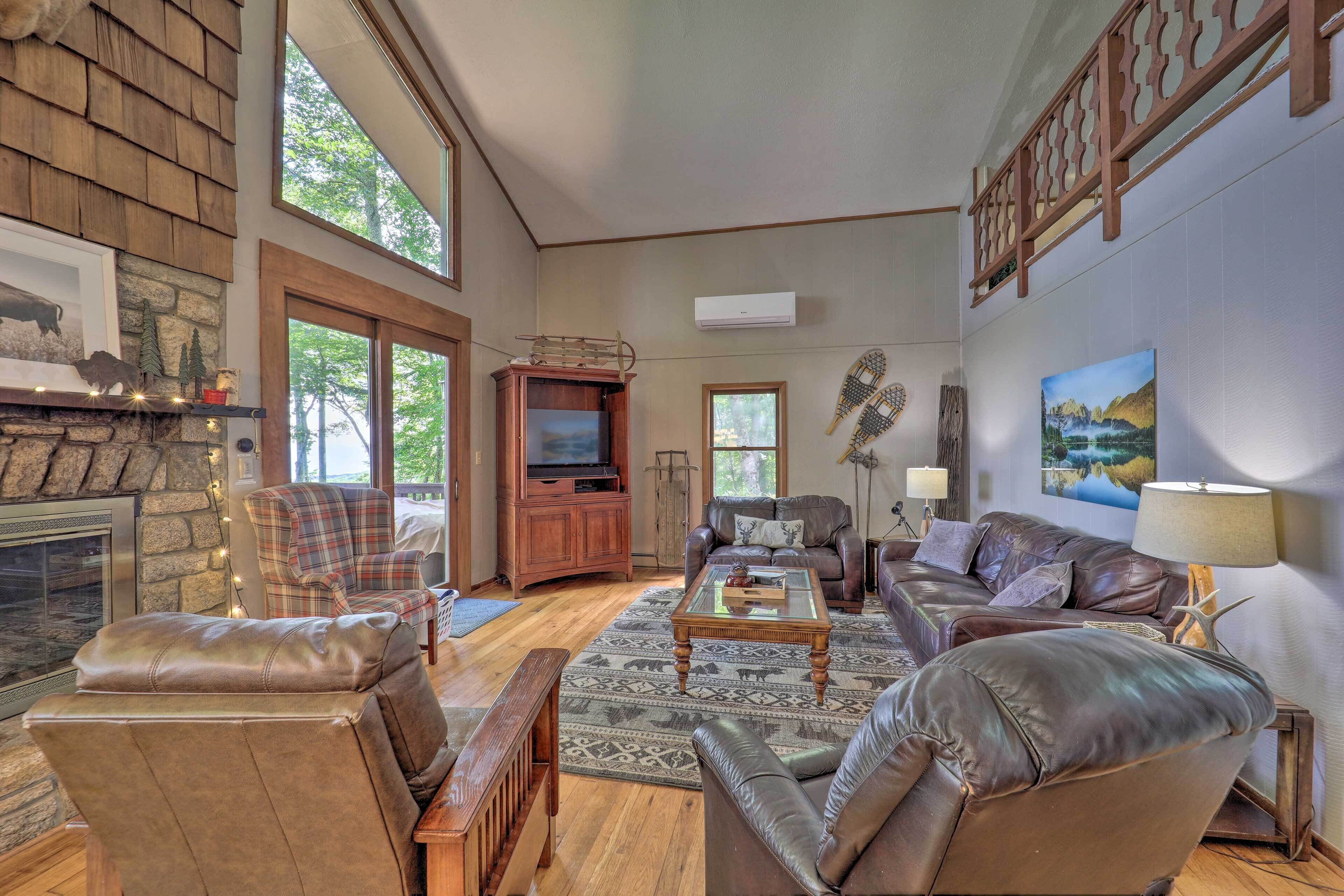 A spacious living room with a stone fireplace, leather sofas, a television, and a large window overlooking greenery.